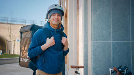 Happy Food Delivery Man Wearing Thermal Backpack on Bike Deliver Restaurant Order. Smiling Courier Looking at Camera. Courier Delivers Takeaway Lunch in Modern City District Office Building.の写真素材