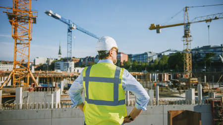 Confident Bearded Head Civil Engineer-Architect in Sunglasses is Standing Outside with His Back to Camera in a Construction Site on a Bright Day. Man is Wearing a Hard Hat, Shirt and a Safety Vest.の写真素材