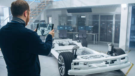 Male Engineer is Working in a High Tech Development Facility Holding a Tablet Computer. He Stands Next to an Electric Car Chassis Prototype with Batteries and Wheels.の写真素材