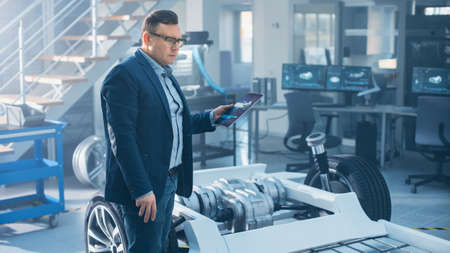 Engineer with Glasses Works on a Tablet Computer Next to an Electric Car Chassis Prototype with...