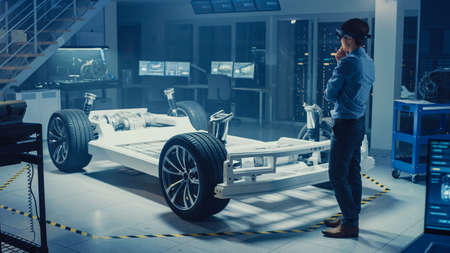 Automotive Engineer Working on Electric Car Chassis Platform, Using Augmented Reality Headset. In Innovation Laboratory Facility Concept Vehicle Frame Includes Wheels, Suspension, Engine and Battery.の写真素材