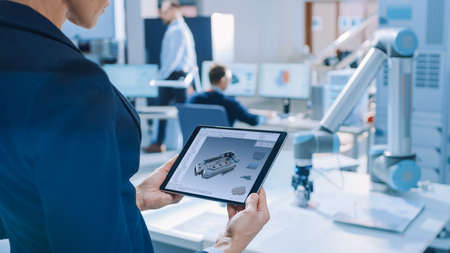 Stylish Female Industrial Robotics Engineer Uses Tablet Computer with CAD Software and to Manipulate and Program Robot Arm for Lifting, Moving Objects. Technology Research Facility for Machineの写真素材