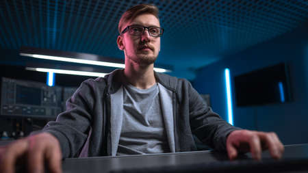 Low Angle Portrait Shot of Software Developer Hacker Wearing Glasses Sitting at His Desk and Working...