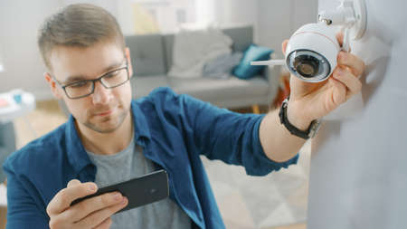 Young Man in Glasses Wearing a Blue Shirt is Adjusting a Modern Wi-Fi Surveillance Camera with Two...