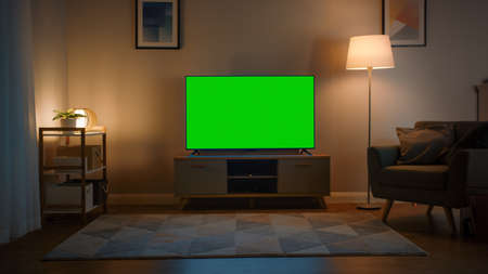 Shot of a TV with Horizontal Green Screen Mock Up. Cozy Evening Living Room with a Chair and Lamps Turned On at Home.の写真素材