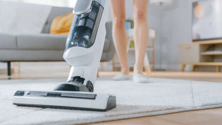 Close Up Shot of a Young Beautiful Woman in Jeans Shirt and Shorts Vacuum Cleaning a Carpet in a Bright Cozy Room at Home. She Uses a Modern Cordless Vacuum. Shes Happy and Cheerful.の写真素材