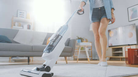 Close Up Shot of a Young Beautiful Woman in Jeans Shirt and Shorts Vacuum Cleaning a Carpet in a Bright Cozy Room at Home. She Uses a Modern Cordless Vacuum. Shes Happy and Cheerful.の写真素材
