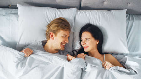 Happy and Fun Young Couple Lying under Blanket on a Bed, Looking at Each other and Smiling. Lovely...