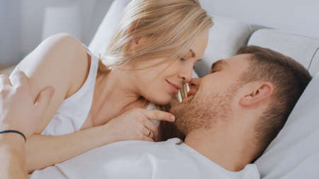 Happy Young Couple Cuddling Together in the Bed, Kissing and Smiling. Beautiful Blonde Female and Her Fit Loving Partner Experiencing Tender and Loving Moments of Living Together.の写真素材