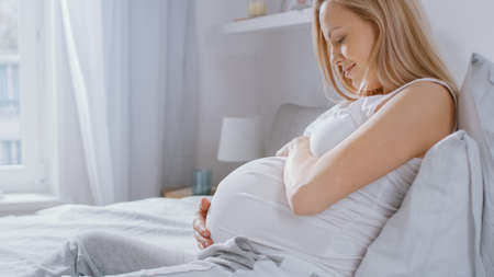 Happy Young Pregnant Woman Laying in Bed in the Morning, She Touches and Caresses Her Belly Tenderly, Loving Her Future Baby and Every Moment of Her Life.の写真素材