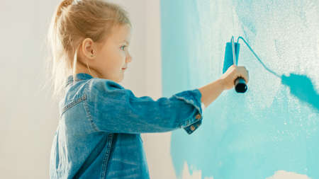Adorable Small Girl in Jeans Coat is Painting a Wall. She Paints with Roller that is Covered in Light Blue Paint. Room Renovations at Home.の写真素材