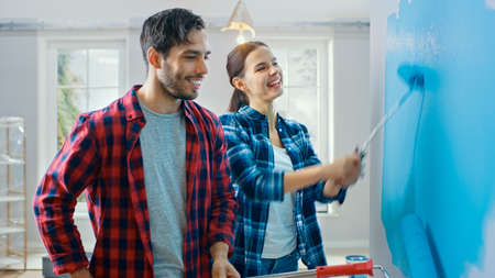 Young Beautiful Couple Talk and Decorate Their New Apartment. Husband and Wife are Painting the Wall with Rollers that are Dipped in Light Blue Paint. Renovations at Home.の写真素材