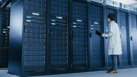 In Data Center: Male IT Specialist Wearing White Coat Stands Beside Server Rack, Uses Laptop...