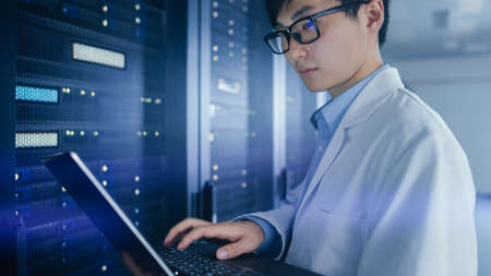 In Data Center: Male IT Specialist Wearing White Coat Stands Beside Server Rack, Uses Laptop...