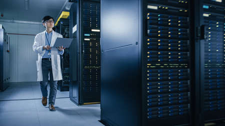 In Data Center: Male IT Technician Wearing White Coat Walking alongside Server Racks, Uses Laptop...