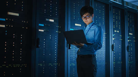 In Dark Data Center: Male IT Specialist Walks along the Row of Operational Server Racks, Uses Laptop...