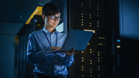 In Dark Data Center: Male IT Specialist Stands Beside the Row of Operational Server Racks, Uses...
