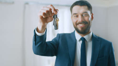 Successful Real Estate Agent in a Suit Smiles and Offers Keys From a New Apartment. Standing in the...