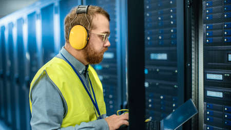 Bearded IT Specialist in Glasses and Headphones, wearing High Visibility Vest is Working on Laptop...
