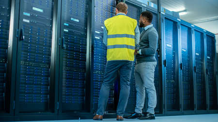 IT Administrator with a Laptop Computer in High Visibility Vest and Black Male Engineer are Talking in Data Center while Standing Before a Server Rack. Running Diagnostics or Doing Maintenance Work.の写真素材