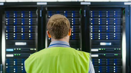 Back Shot of an IT Administrator in High Visibility Vest Walking Towards a Server Rack in Data...