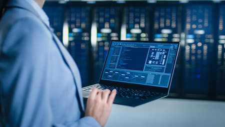 Back View Close Up Shot of an IT Specialist Working on Laptop in Data Center Next to Server Racks....