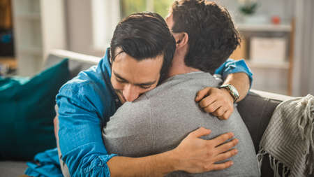 Cute Attractive Male Gay Couple Sit Together on a Sofa at Home. Boyfriends are Hugging and Embracing Each Other. They are Happy and Smiling. They are Casually Dressed and Room Has Modern Interior.の写真素材