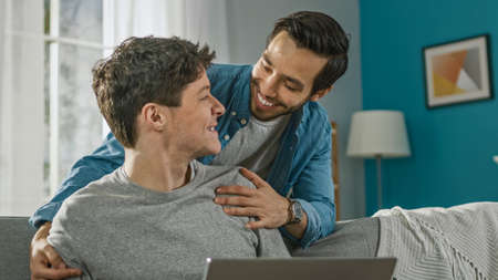 Sweet Male Gay Couple Spend Time at Home. Young Man Works on a Laptop, His Partner Comes From Behind...