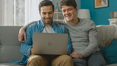 Adorable Male Gay Couple Spend Time at Home. They Sit on a Sofa and Use the Laptop. They Browse Online. Partner Puts His Hand Around His Lover. Room Has Modern Interior.の写真素材