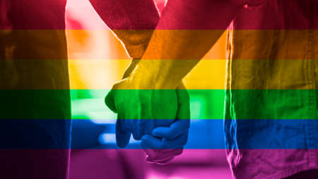Close-Up of Two Male Hands Gently Touching and Hold Each Other. Cute Queer Relationship Concept. Gay Couple are Casually Dressed. Room is Bright and Sunny. Shot has Freedom Rainbow Flag Filter on itの写真素材