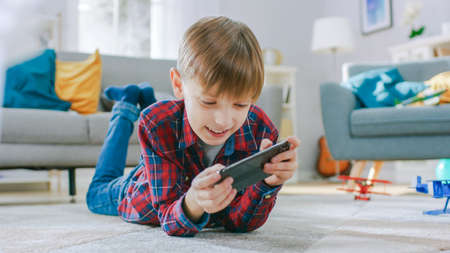 Smart Little Boy Laying on a Carpet Plays in Video Game on His Smartphone, Holds Mobile Phone in Horizontal Landscape Mode. Child Has Fun Playing Videogame in Sunny Living Room.の写真素材