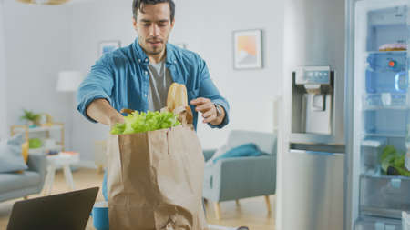 Handsome Attractive Young Man Brings Groceries to the Kitchen. He Bought Fresh Salad Greens. Modern Fridge is on the Background. Room Has Bright Modern Interior.の写真素材