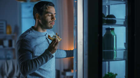 At Night in the Kitchen Handsome Attractive Young Man is Eating a Leftover Pizza from the Fridge. He is Hungry and Feels Satisfied.の写真素材