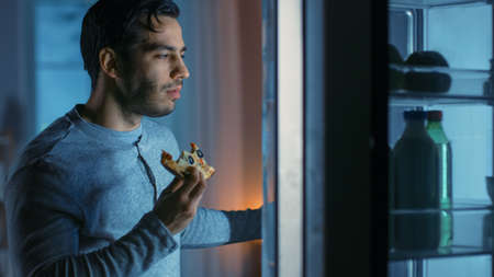At Night in the Kitchen Handsome Attractive Young Man is Eating a Leftover Pizza from the Fridge. He is Hungry and Feels Satisfied.の写真素材