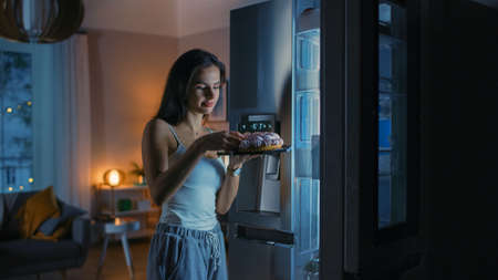Hungry Beautiful Young Woman in the Kitchen in the Evening Opened the Fridge, Takes out a Piece of a Delicious Creamy Cake and Starting to East it. She Feels Satisfied.の写真素材
