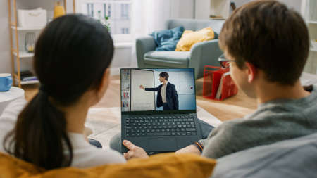 Young Man and Woman at Home Watching Online Lesson on Laptop Computer. They are Sitting on Couch in Living Room. Couple in Love Talking and Watching TV Programme. Friendly Teacher Explains Lesson.の写真素材