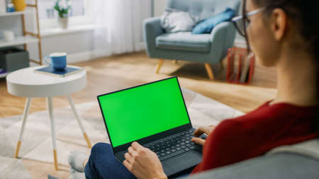 Young Woman at Home Works on a Laptop Computer with Green Mock-up Screen. Shes Sitting On a Couch in His Cozy Living Room. Over the Shoulder Shotの写真素材