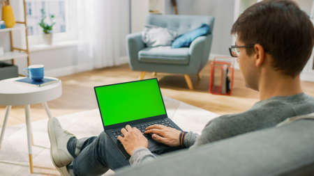 Young Man at Home Works on a Laptop Computer with Green Mock-up Screen. Hes Sitting On a Couch in His Cozy Living Room. Over the Shoulder Shot.の写真素材