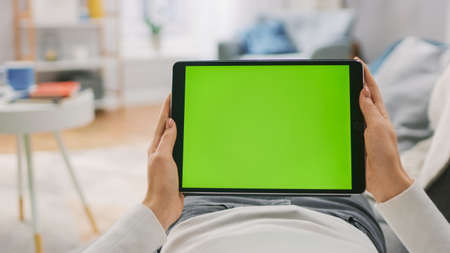 Young Woman at Home Laying on a Couch Using with Green Mock-up Screen Tablet Computer in Horizontal Landscape Mode. Girl Using Touchscreen Device, Browsing Internet, Watching Content, Videos, Blogs.の写真素材