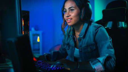 Pretty and Excited Black Gamer Girl in Headphones is Playing First-Person Shooter Online Video Game on Her Computer. Room and PC have Colorful Neon Led Lights. Cozy Evening at Home.の写真素材