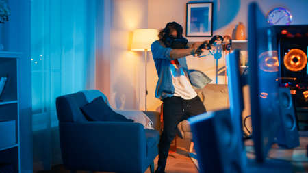 Young Man is Energetically Playing a Virtual Reality Video Game While Wearing a Headset. Hes is Standing in a Cozy Room with Warm Neon Lights.の写真素材