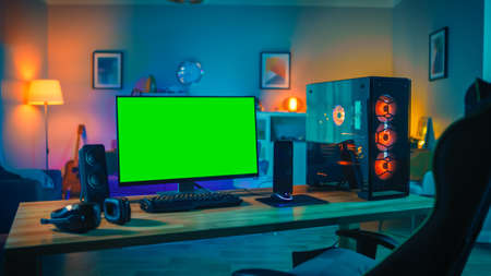 Powerful Personal Computer Gamer Rig with Mock Up Green Screen Monitor Stands on the Table at Home. Cozy Room with Modern Design is Lit with Warm and Neon Light.の写真素材