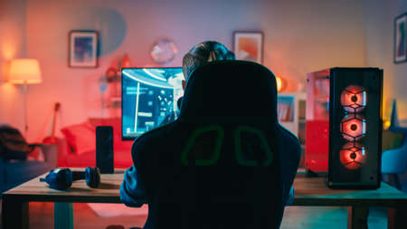 Back Shot of a Gamer Playing First-Person Shooter Online Video Game on His Powerful Personal Computer. Room and PC have Warm Colorful Neon Led Lights. Cozy Evening at Home.の写真素材