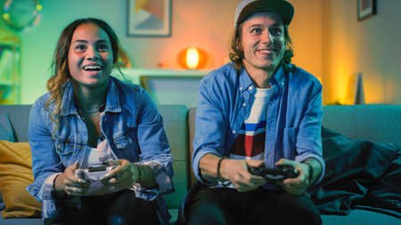 Excited Black Gamer Girl and Young Man Sitting on a Couch and Playing Video Games on Console. They...