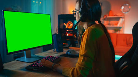 Excited and Concentrated Gamer Girl in Glasses and Headset with a Mic Playing on Her Personal Computer with Green Screen Mock-up Template. Colorful Warm Neon Led Lights. Cozy Evening at Home.の写真素材