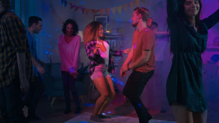 At the College House Party: Diverse Group of Friends Have Fun, Dancing and Socializing. Boys and Girls Dance in the Living Room. Disco Neon Strobe Lights Illuminating Room.の写真素材