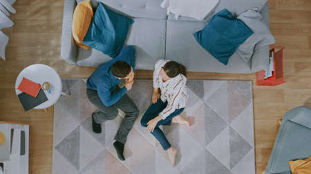 Young Couple is Sitting on a Floor and Talking. They Have an Emotional Dialogue. Cozy Living Room with Modern Interior with Carpet, Sofa, Chair, Table, Book Shelf, Plant and Wooden Floor. Top Down.の写真素材