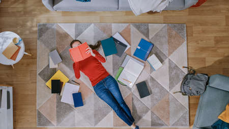 Young Girl in Red Coat and Blue Jeans is Lying Down on a Floor, Reading a Notebook. Looks Above, Smiles and Laughs. Cozy Living Room with Modern Interior with Carpet, Workbooks and Backpack. Top View.の写真素材