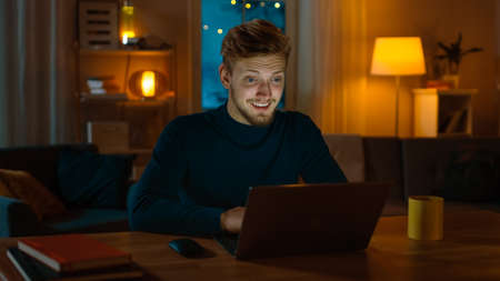 Handsome Smiling Man Works on a Laptop while Sitting at His Desk at Home. Young Freelancer Works on Computer in His Cozy Living Room with Warm Evening Lighting Turned on.の写真素材