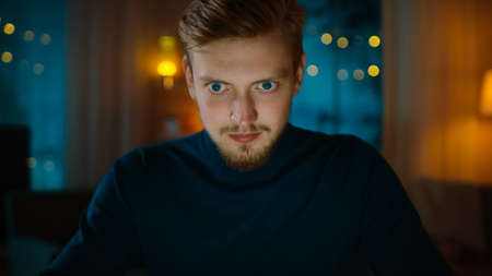 In the Evening: Man Sitting at His Desk and Using Laptop. He is Nervous and Fully Concentrated on His Important Job. In the Background Cozy Living Room.の写真素材